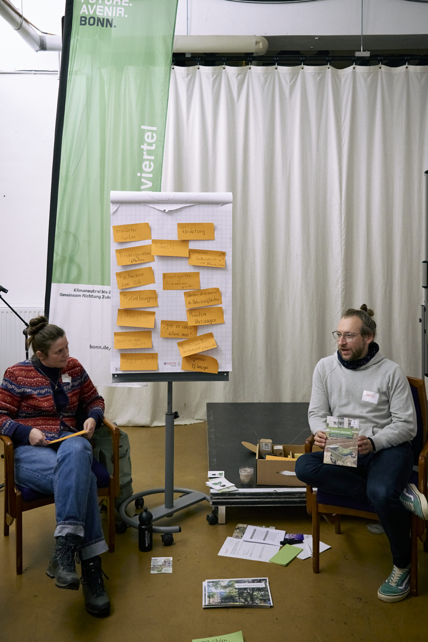 Zwei Workshopleitende sitzen mit Moderationsmaterial und einem Flipchart im Raum. Auf dem Flipchart sind zahlreiche beschriebene Moderationskarten zu sehen.